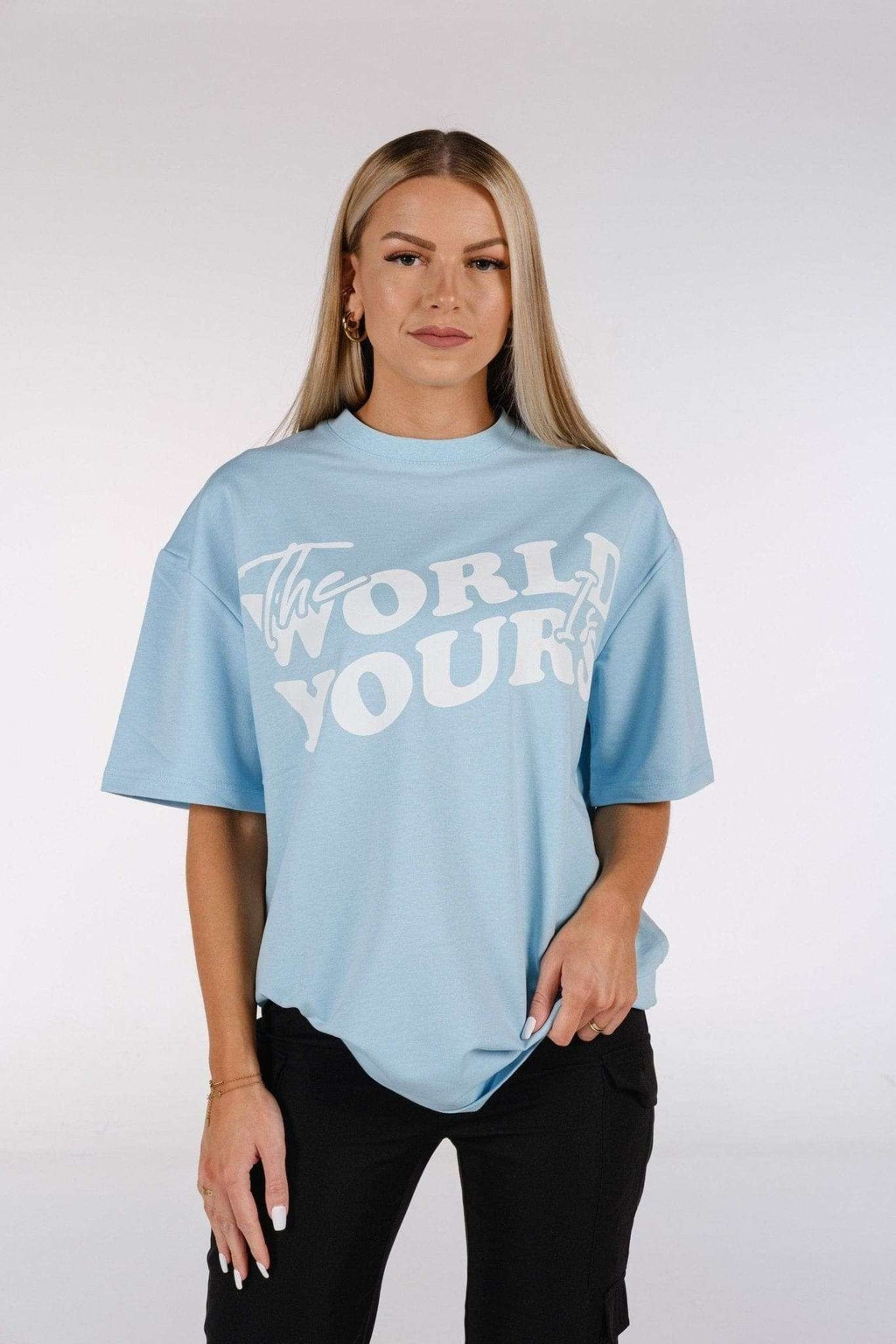 1. Woman wearing baby blue oversized t-shirt with 'The World Is Yours' print, standing in studio setting