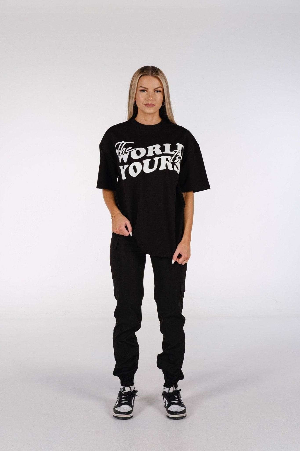 2. Woman in oversized black t-shirt with 'The World Is Yours' print, paired with black pants and sneakers, standing in studio