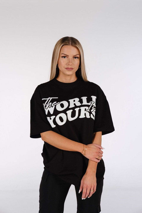1. Woman wearing oversized black t-shirt with 'The World Is Yours' print, standing with arms crossed in studio setting