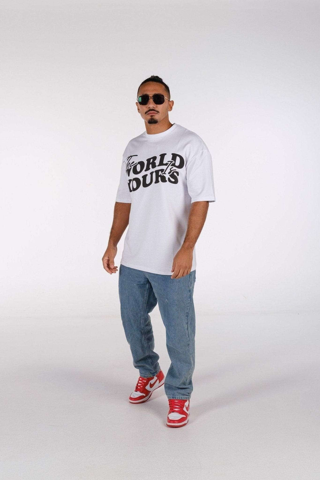 3. Man in white oversized t-shirt with 'The World Is Yours' print, wearing jeans and red sneakers in studio