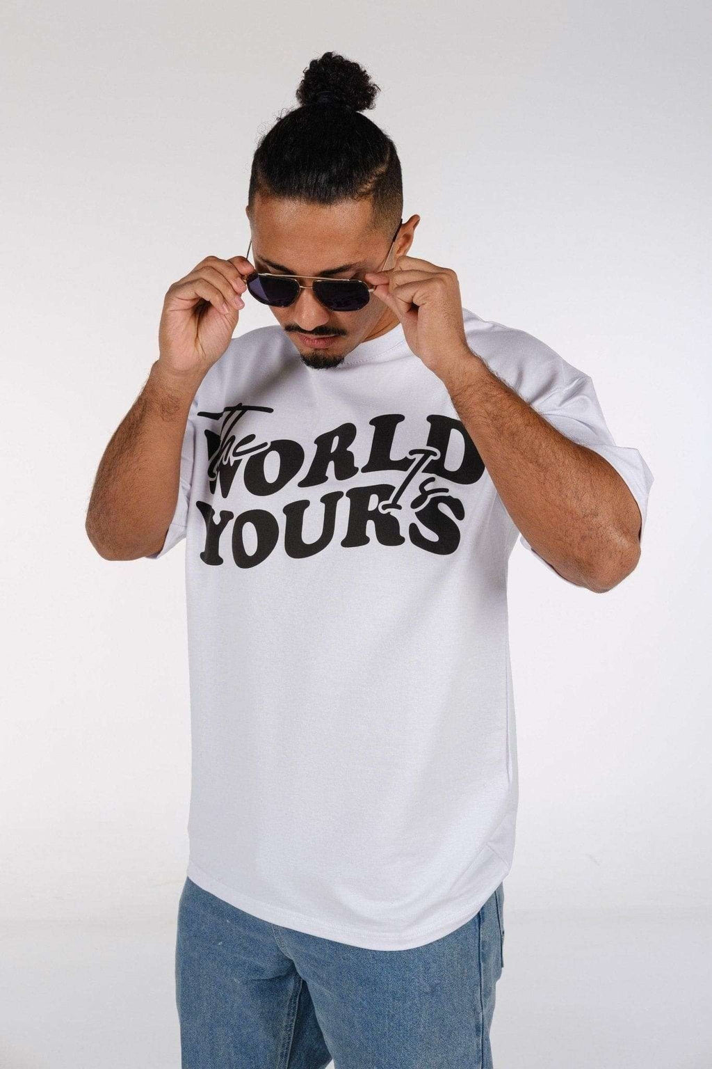 2. Man wearing white oversized t-shirt with 'The World Is Yours' print, adjusting sunglasses in studio