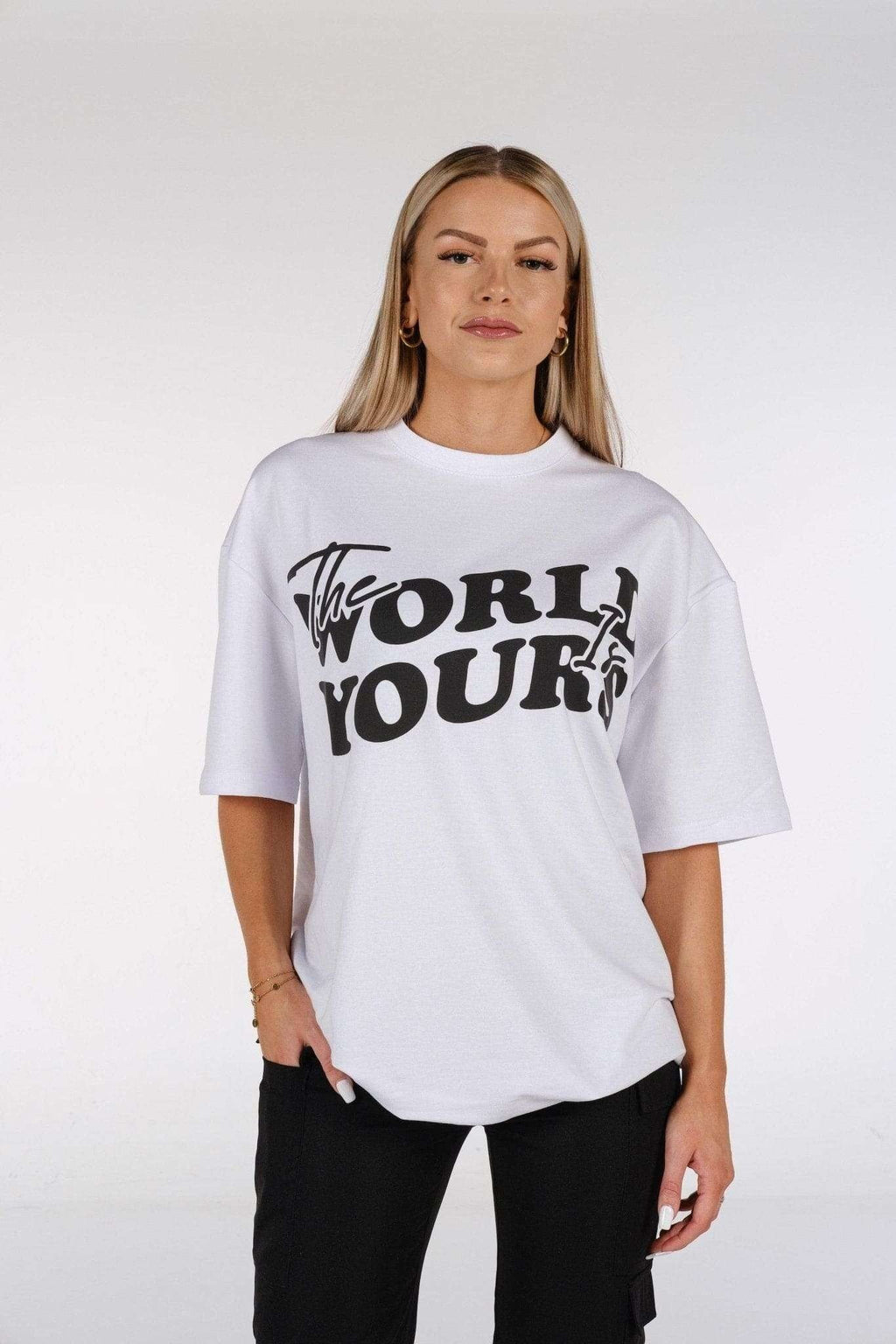 1. Woman wearing white oversized t-shirt with 'The World Is Yours' print, standing in studio setting