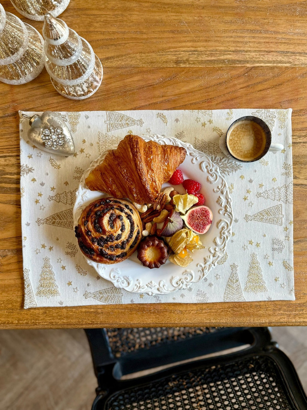 1. Lifestyle image of Hortensias Home Golden Forest table placemat with breakfast setting including croissant, pastry, and coffee on a wooden table