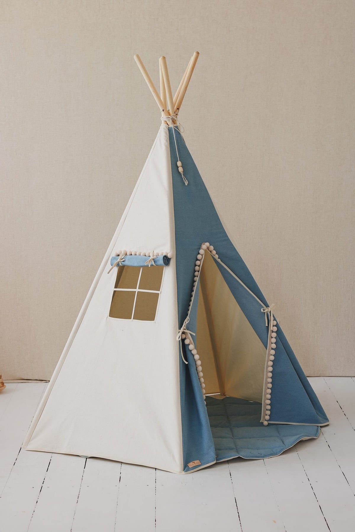 6. Side view of blue and white children's teepee tent with pompoms, open entrance, on white floor against beige wall.
