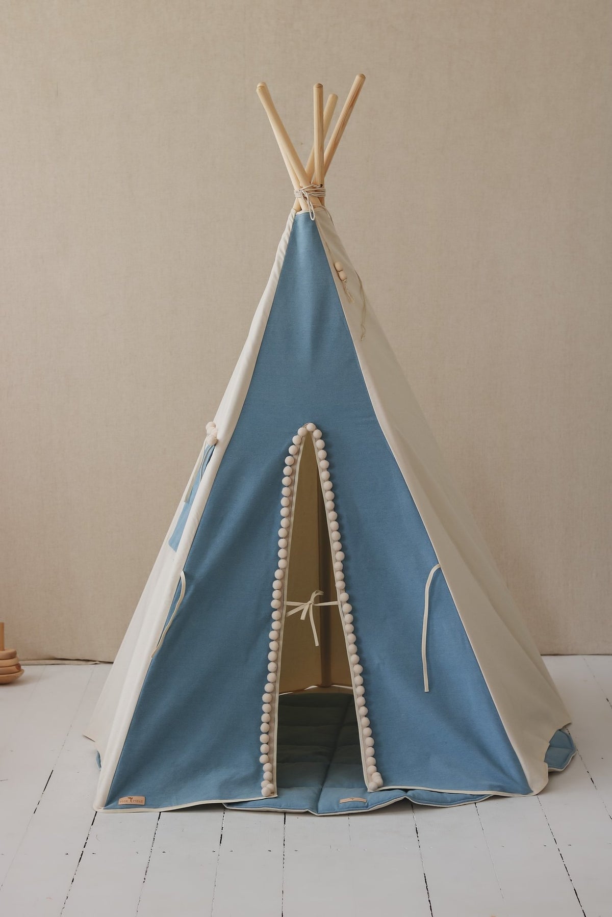 2. Front view of blue and white children's teepee tent with pompoms, closed entrance, on white floor against beige wall.