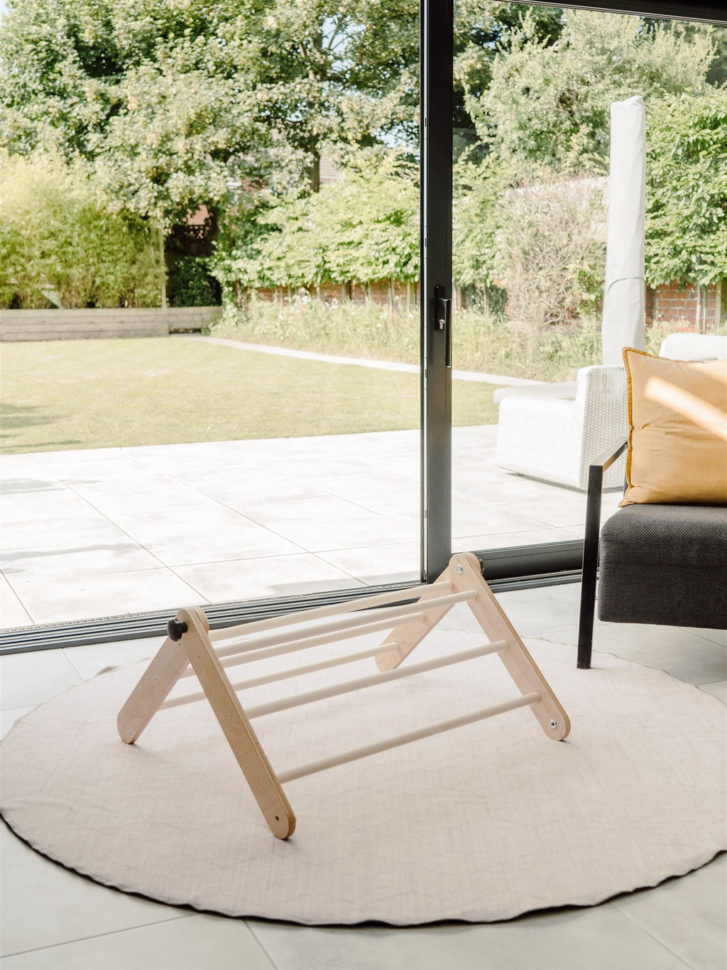 3. Family-scl Pikler Triangle in birch plywood placed on a round rug in a bright room with garden view, emphasizing its natural material and home integration.