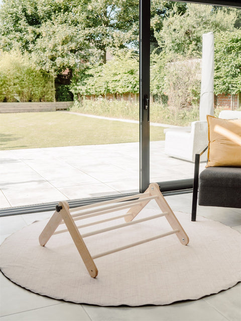 3. Family-scl Pikler Triangle in birch plywood placed on a round rug in a bright room with garden view, emphasizing its natural material and home integration.