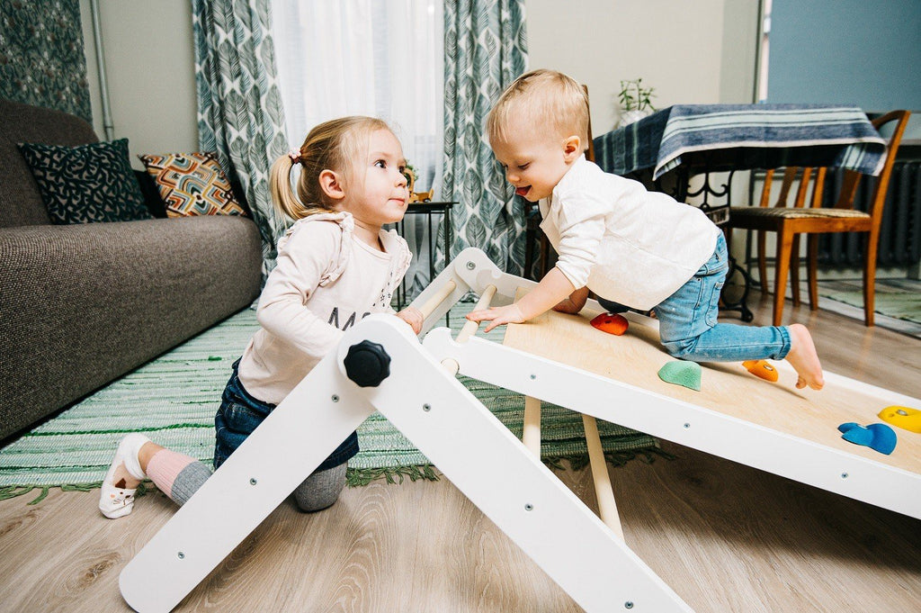 1. Two children playing on a Family-scl Pikler Triangle with climbing ramp in a living room setting, showcasing its use for physical development.