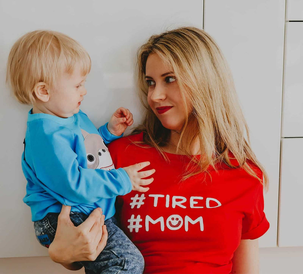 1. Woman wearing red HILP Tired Mom T-shirt holding a child, showcasing casual style