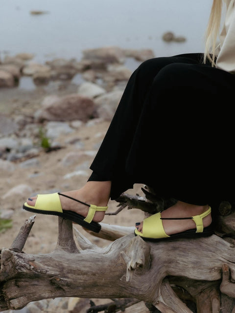 1. Women's TOKU Berlin Sandals in lime yellow worn by model sitting on driftwood by the beach
