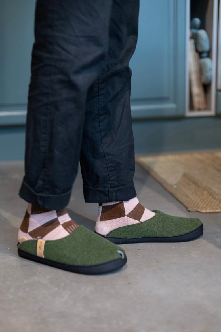 1. Unisex TOKU Brussels Slippers in moss green worn by a person with patterned socks, showcasing comfort and style in a home setting