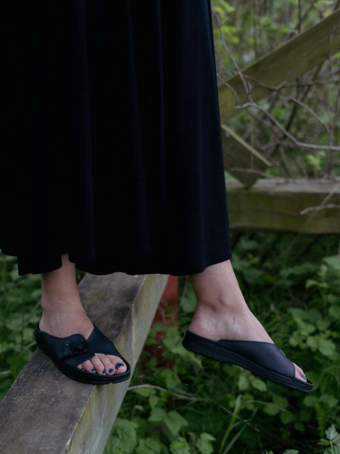 2. Women's black TOKU Rome Sandals by Omaking styled with a long black dress, shown outdoors on a wooden beam