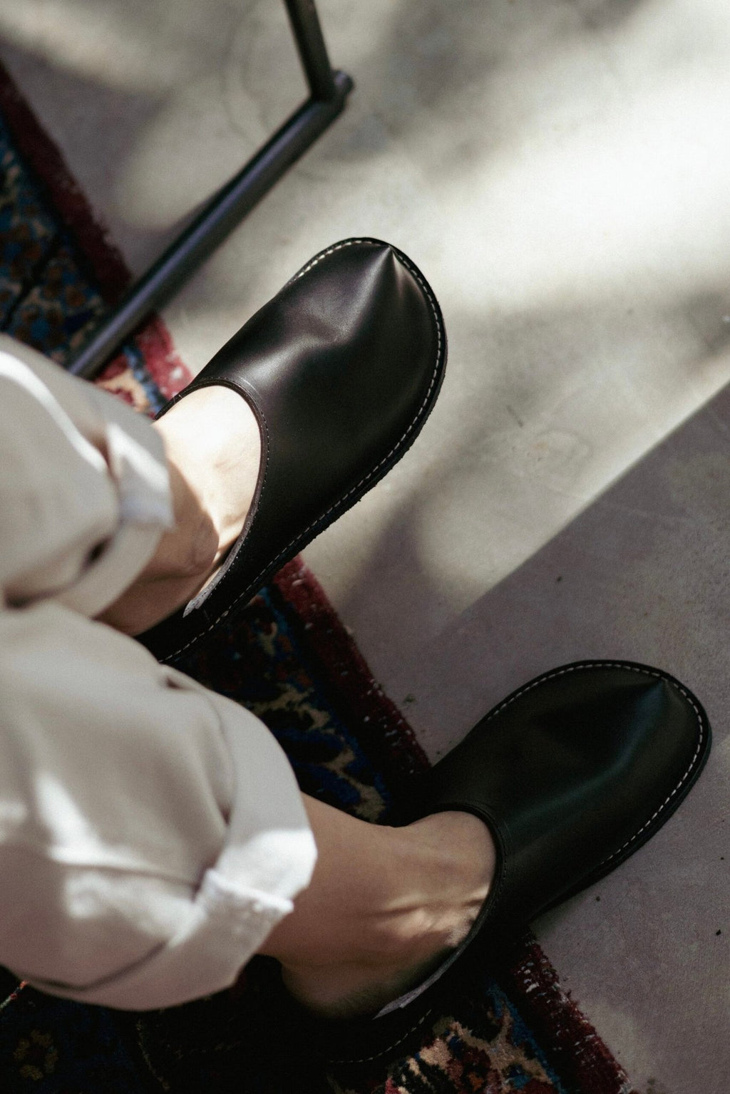1. Person wearing unisex black slip-on shoes with minimalist design, seated on a patterned rug in natural light