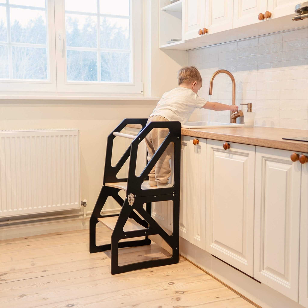 1. Toddler using black kitchen tower at sink in bright kitchen