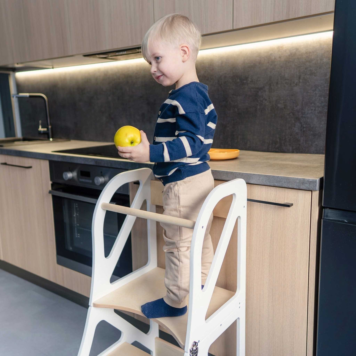 1. Child standing on white transformable kitchen tower holding an apple in a kitchen