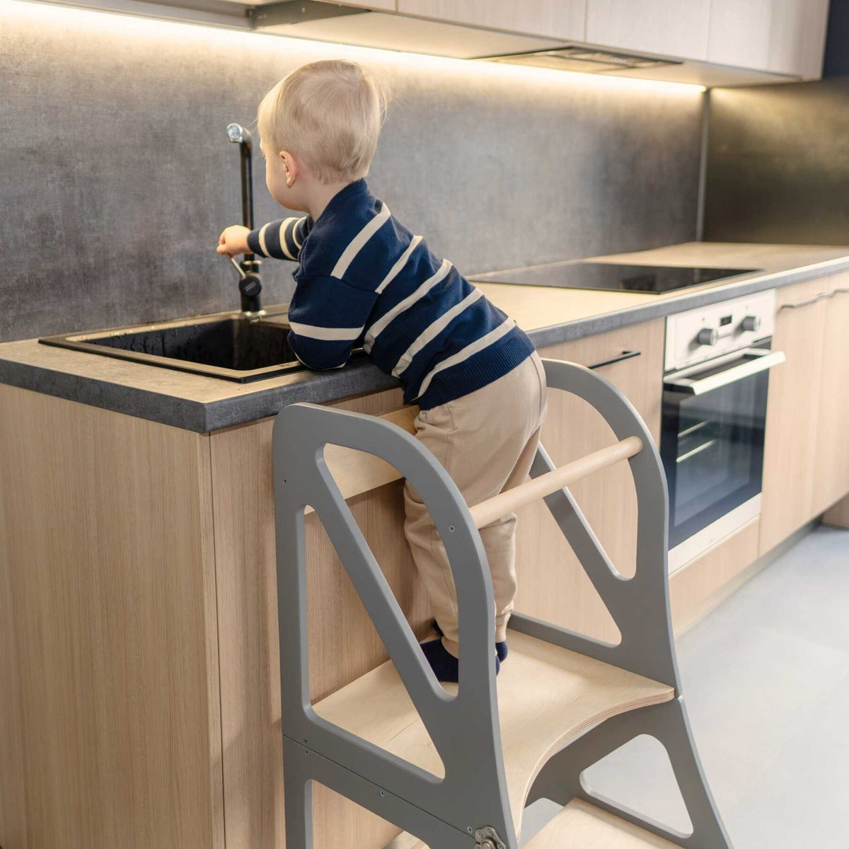 1. Child using gray transformable kitchen tower to reach a kitchen sink