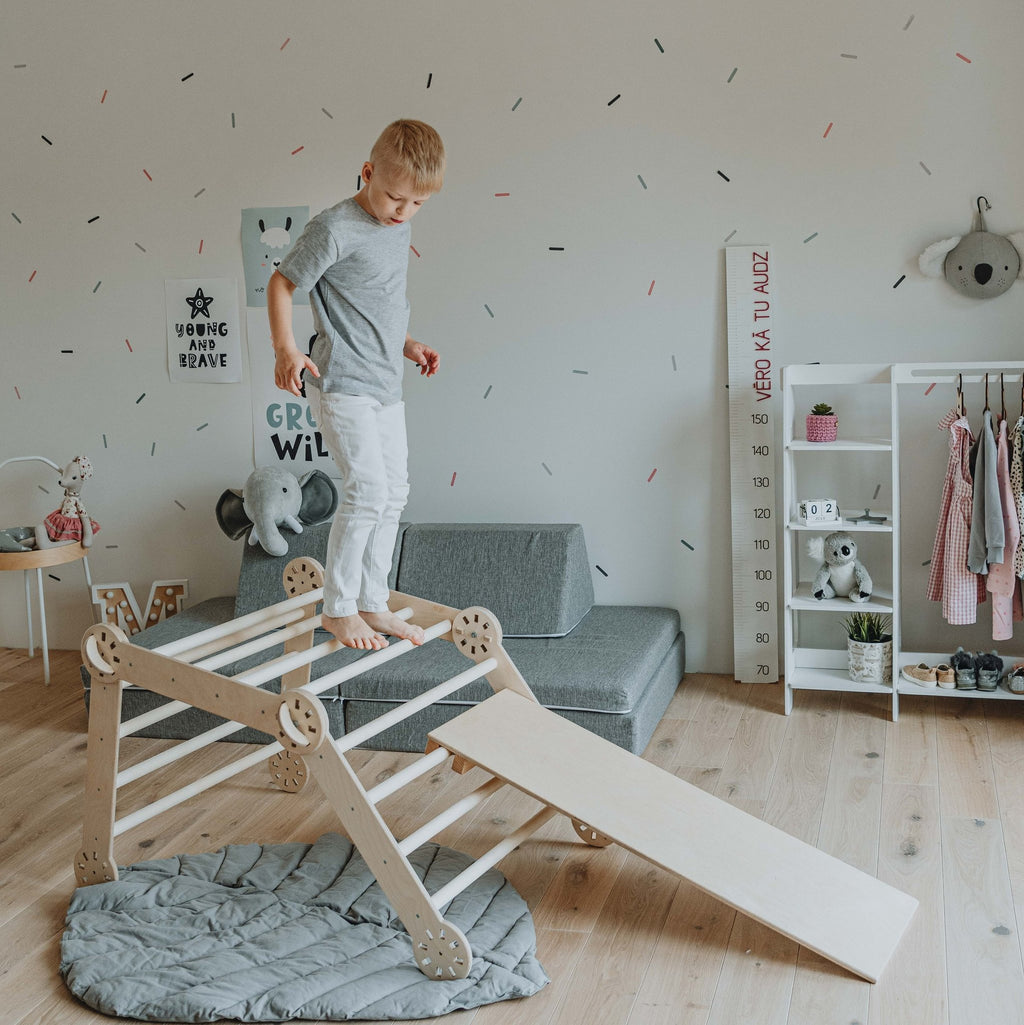 5. Young boy in grey shirt and white pants standing on top of wooden Montessori climber with ramp in a decorated playroom