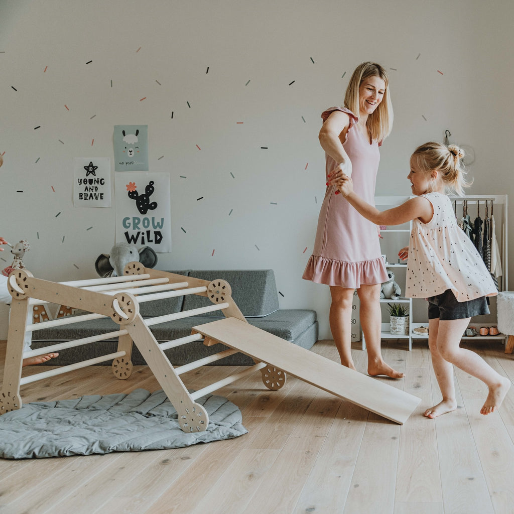4. Woman in pink dress holding hands with young girl on wooden Montessori climber with ramp in a playroom