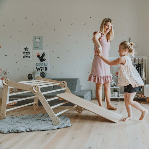 4. Woman in pink dress holding hands with young girl on wooden Montessori climber with ramp in a playroom