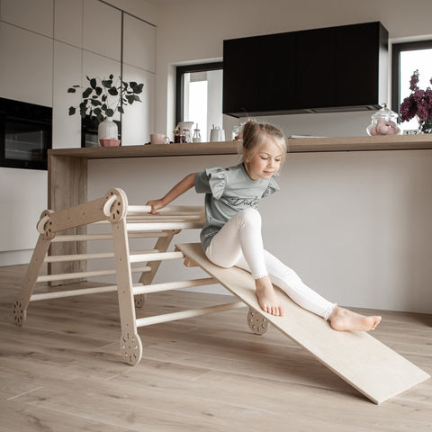 1. Young girl in grey shirt and white leggings sliding down a wooden Montessori climber ramp in a modern kitchen setting