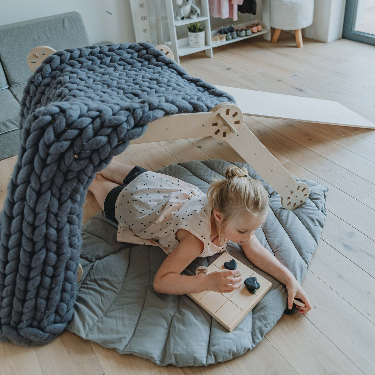 3. Young girl playing under a wooden Montessori climber covered with a blue blanket, lying on a grey mat