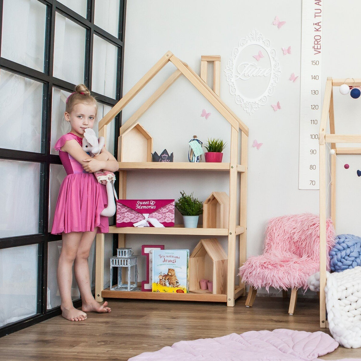 1. Girl in pink dress standing next to natural wood house-shaped shelf with toys