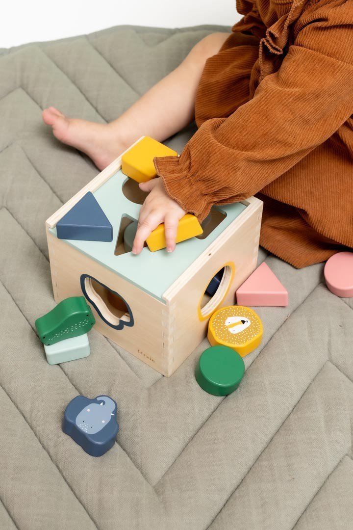 1. Child playing with Trixie Baby wooden shape sorter on quilted mat, featuring colorful geometric shapes and animal designs