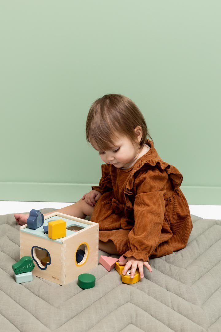 4. Child in brown dress playing with Trixie Baby wooden shape sorter against green wall