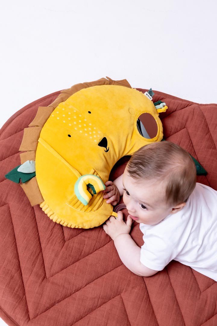 1. Baby on tummy on a brown mat, engaging with Trixie Baby's Mr. Lion tummy time pillow, showing sensory features and lion face design