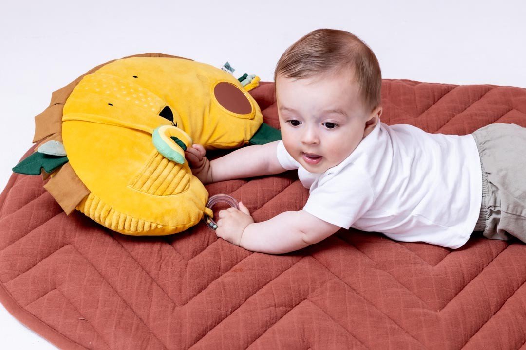 1. Baby lying on tummy on a brown mat, interacting with Trixie Baby's Mr. Lion tummy time pillow, featuring sensory elements and a lion face design