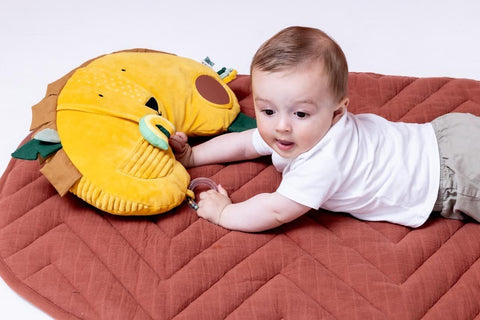1. Baby lying on tummy on a brown mat, interacting with Trixie Baby's Mr. Lion tummy time pillow, featuring sensory elements and a lion face design