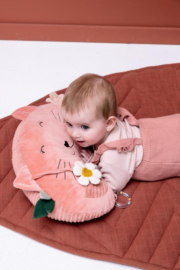 1. Baby lying on pink Trixie Baby tummy time pillow with cat design, featuring sensory elements like a flower and leaf, on a quilted mat