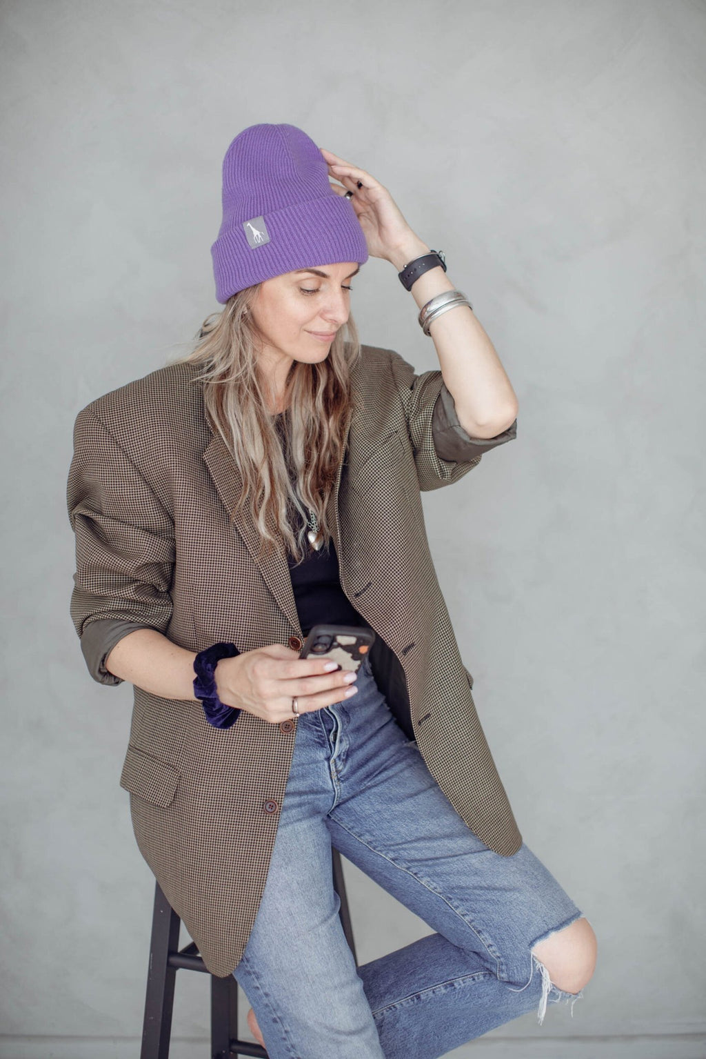 1. Woman wearing purple beanie with giraffe label, sitting on stool in casual outfit, holding phone
