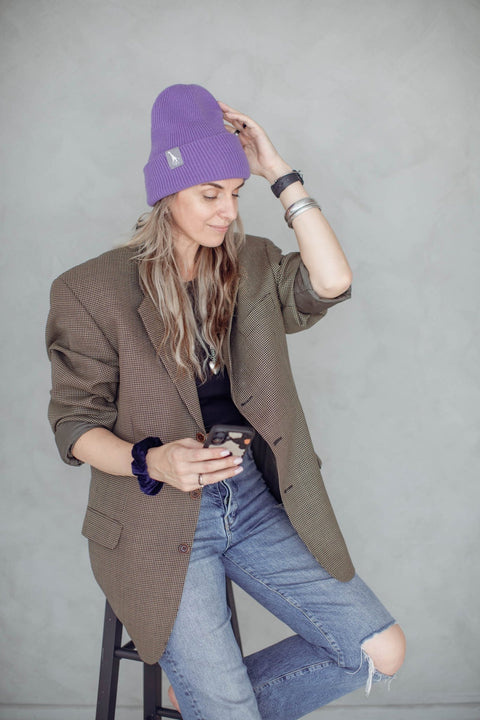1. Woman wearing purple beanie with giraffe label, sitting on stool in casual outfit, holding phone