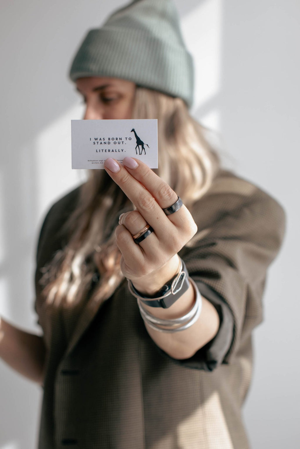 7. Woman holding card with giraffe logo, wearing light green beanie, casual blazer in bright setting