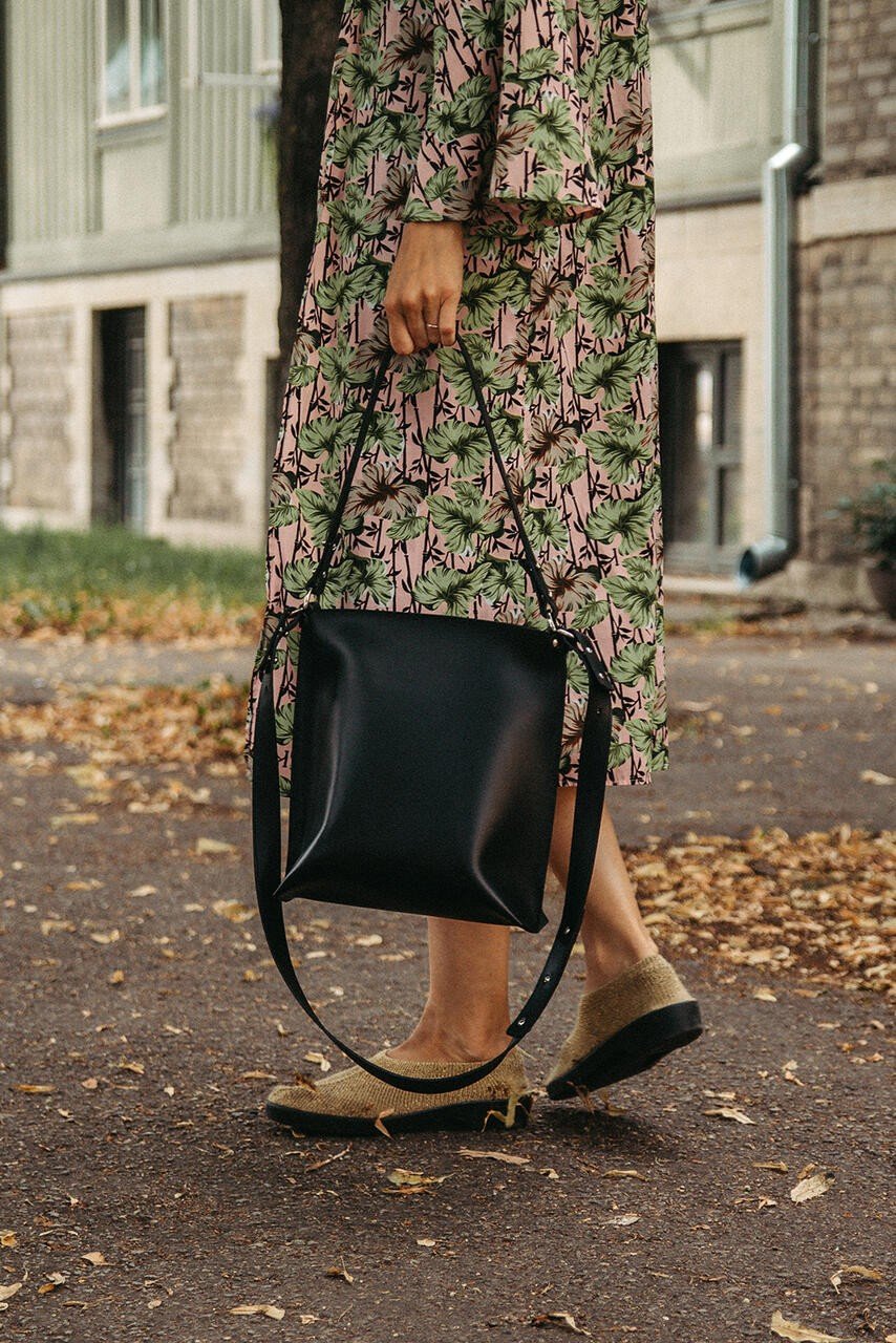 2. Close-up of black Stella Soomlais Escape Artist handbag held by woman in floral dress on leaf-strewn path