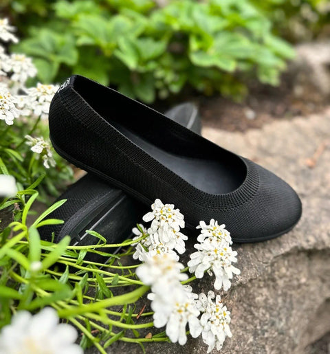 2. Black upcycled ballerina shoes by Kira Sustainable placed on rocks with white flowers, showcasing eco-friendly design
