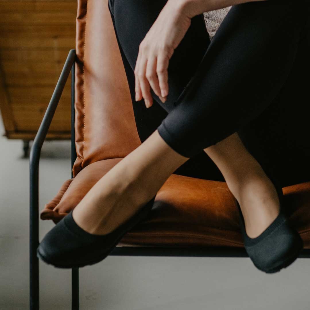 3. Women's black upcycled ballerina shoes by Kira Sustainable, worn by a model sitting on a chair in a cozy setting