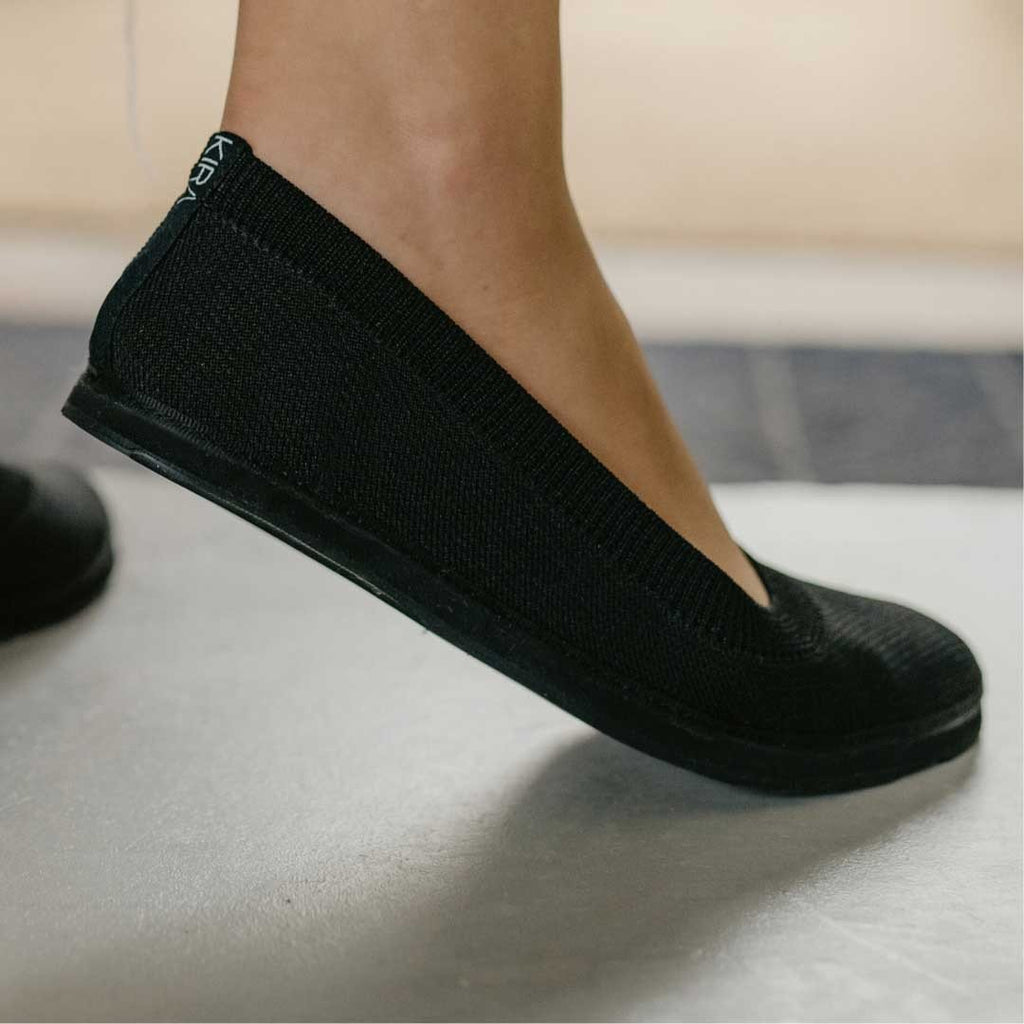 1. Women's black upcycled ballerina shoes by Kira Sustainable, shown in a lifestyle setting with a model wearing them indoors
