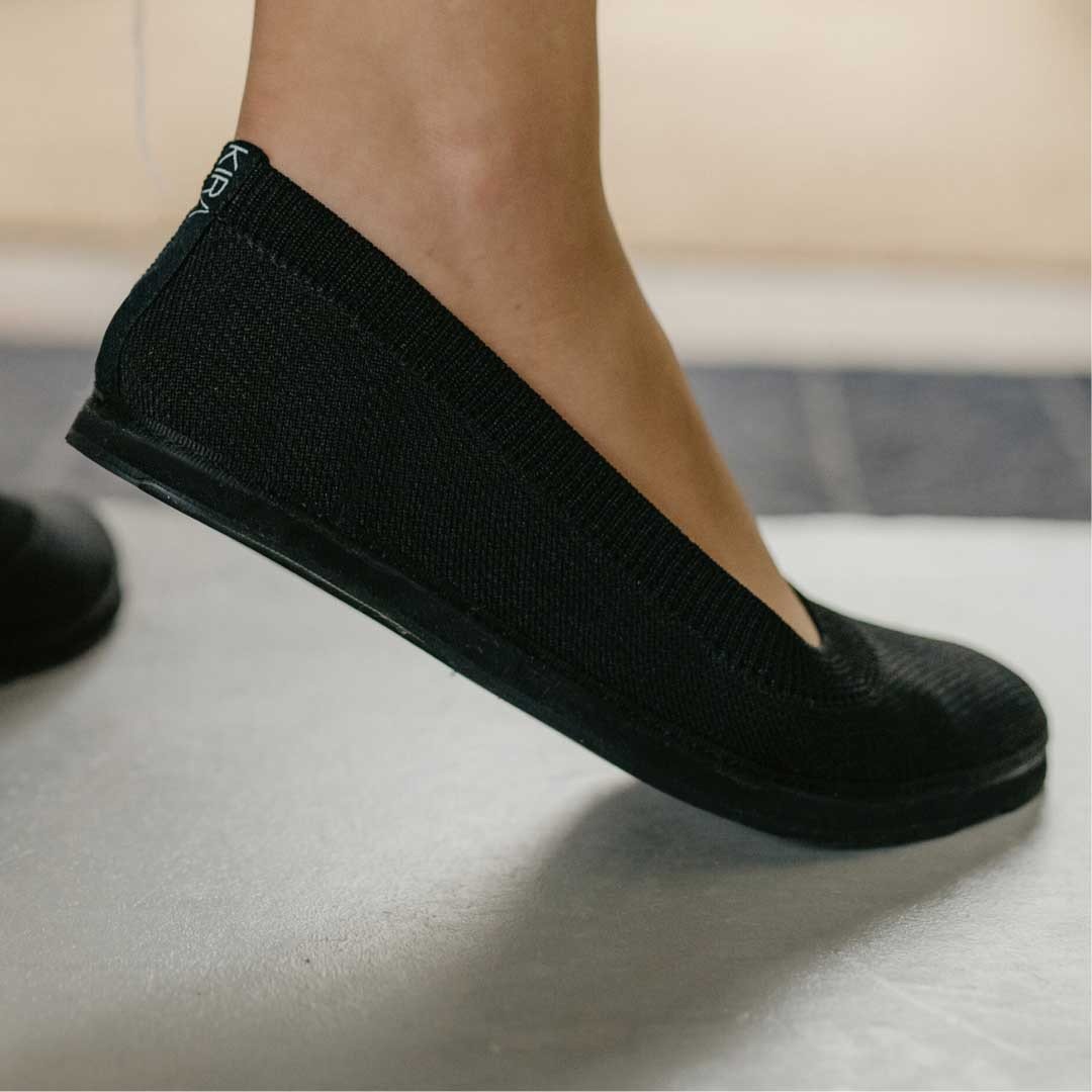 1. Women's black upcycled ballerina shoes by Kira Sustainable, shown in a lifestyle setting with a model wearing them indoors