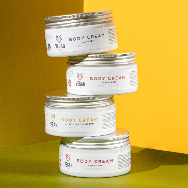6. Stack of Vegan Fox Body Cream jars in various scents including grapefruit, lavender, linden-tree blossom, and red grape on a yellow background