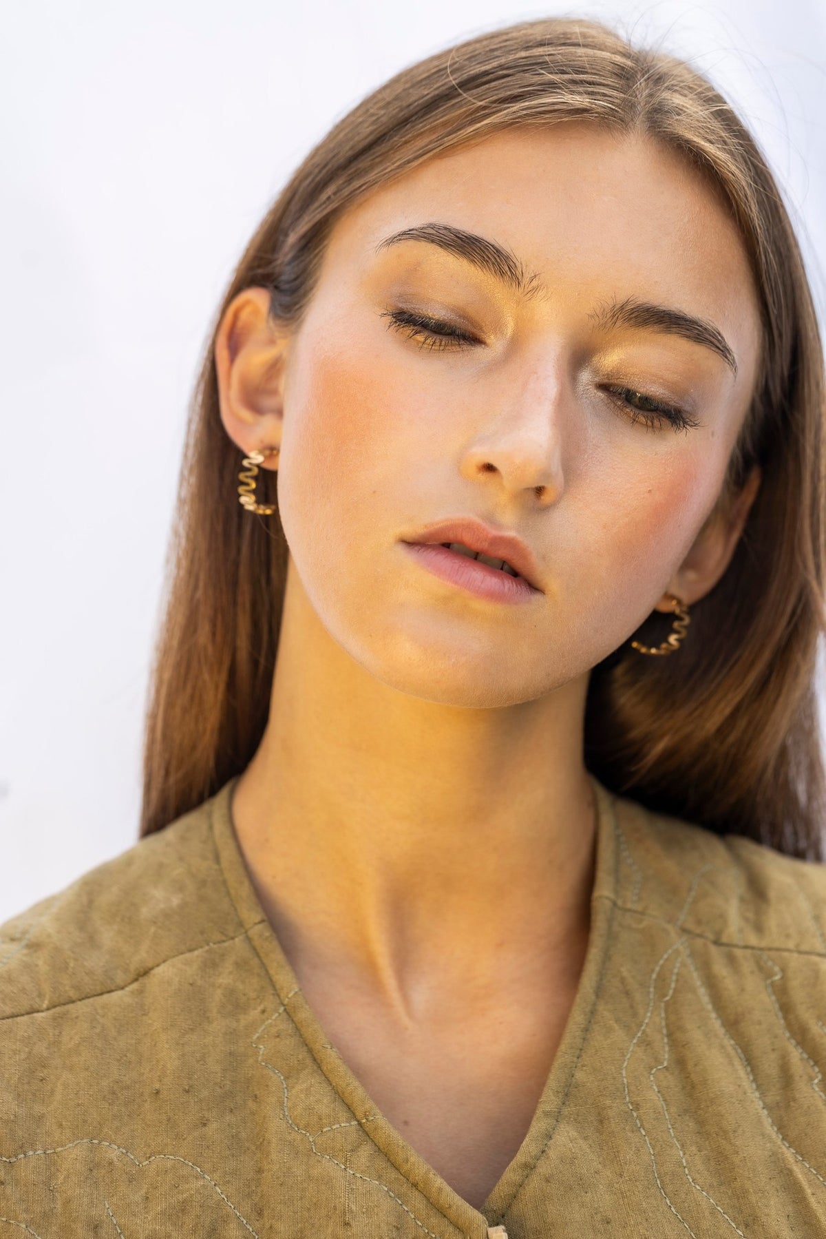 1. Woman wearing Lentsius Vena Sunlit gold-tone wavy earrings, eyes closed with hair down, against a neutral background