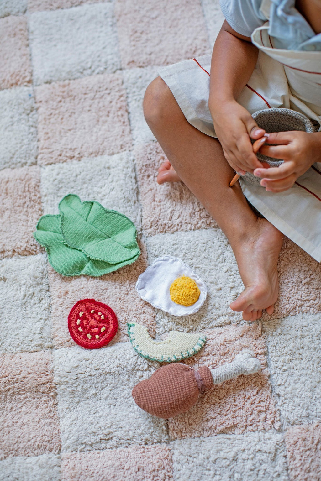 1. Child sitting on Lorena Canals rose and white checkerboard rug with toy food items
