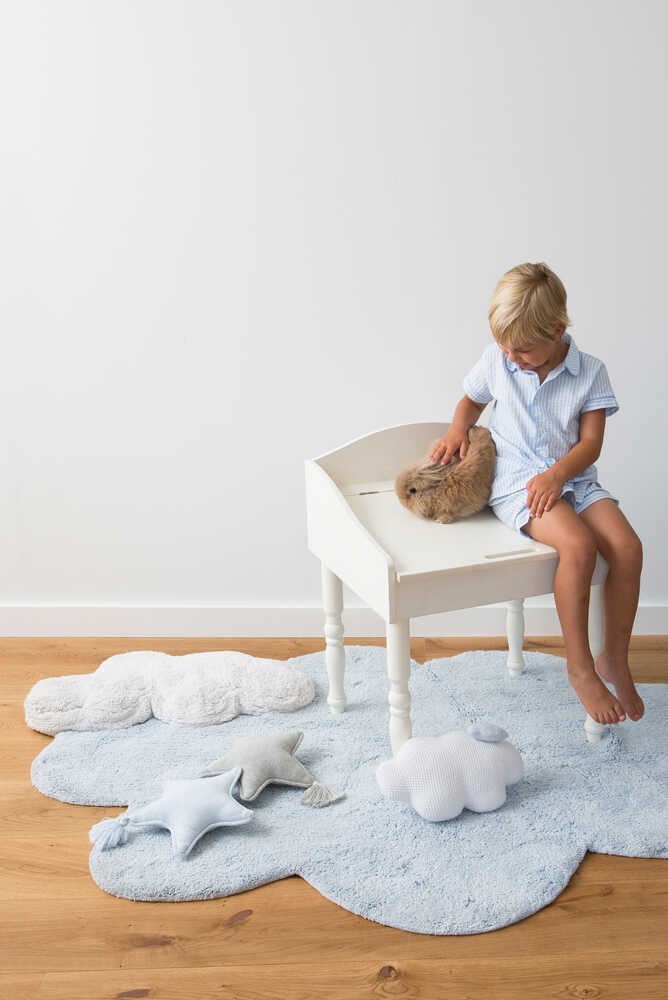 1. Child sitting on a white chair with a rabbit, on a blue cloud-shaped rug with star and cloud pillows in a bright room