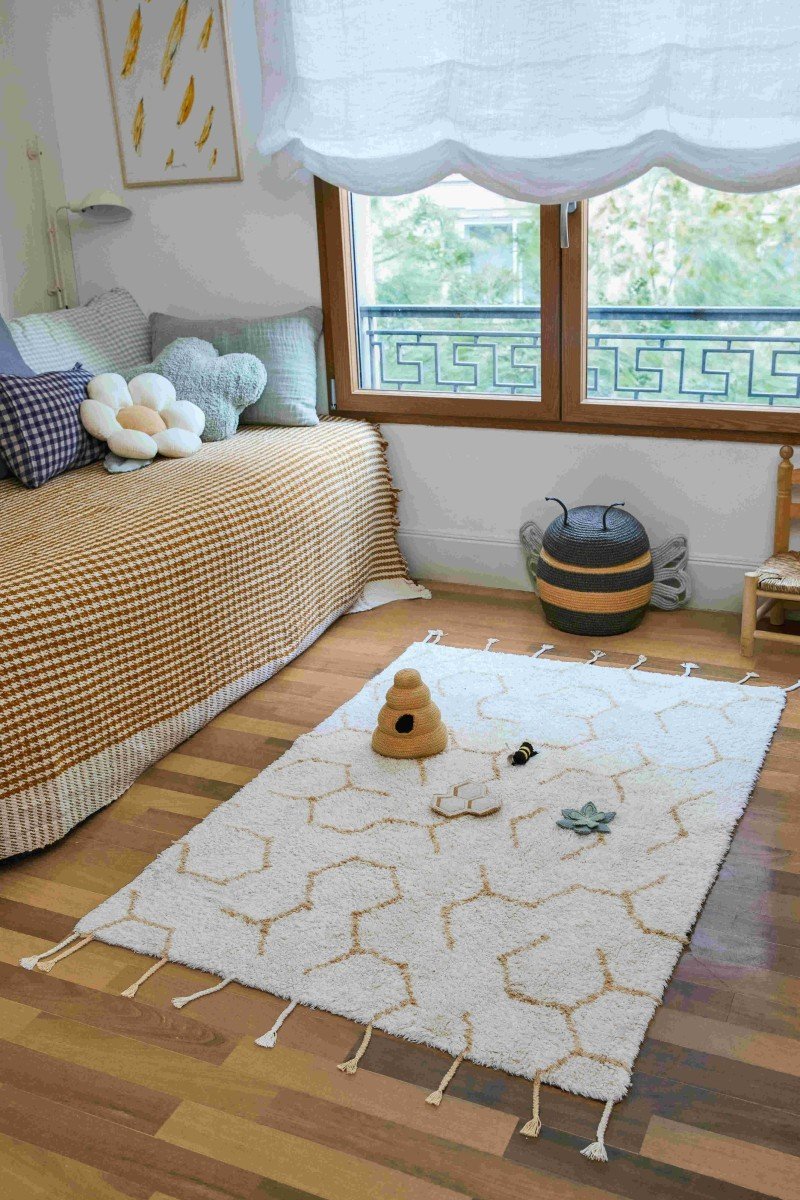 6. Ivory play rug with honeycomb pattern and textile accessories in a child's room with wooden floor and window view