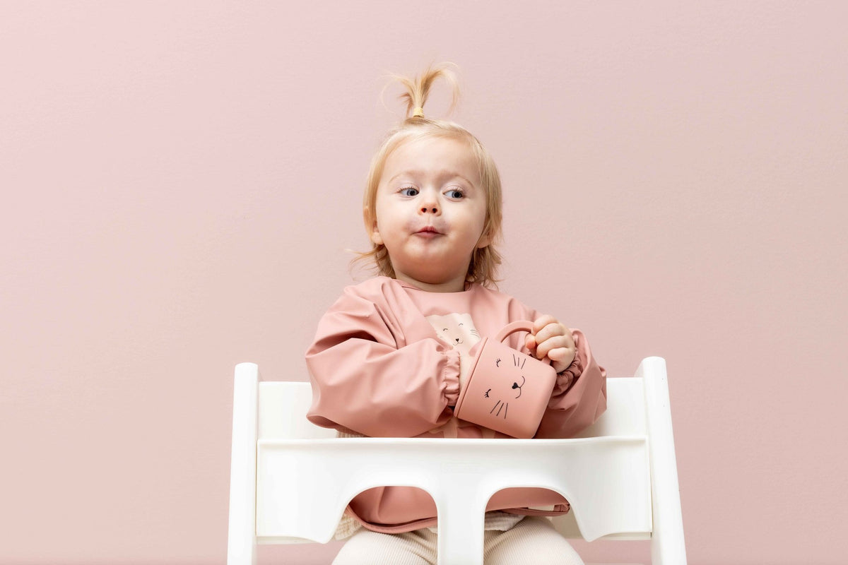 1. Toddler wearing pink waterproof long sleeve bib with cat print, sitting in high chair against pink background