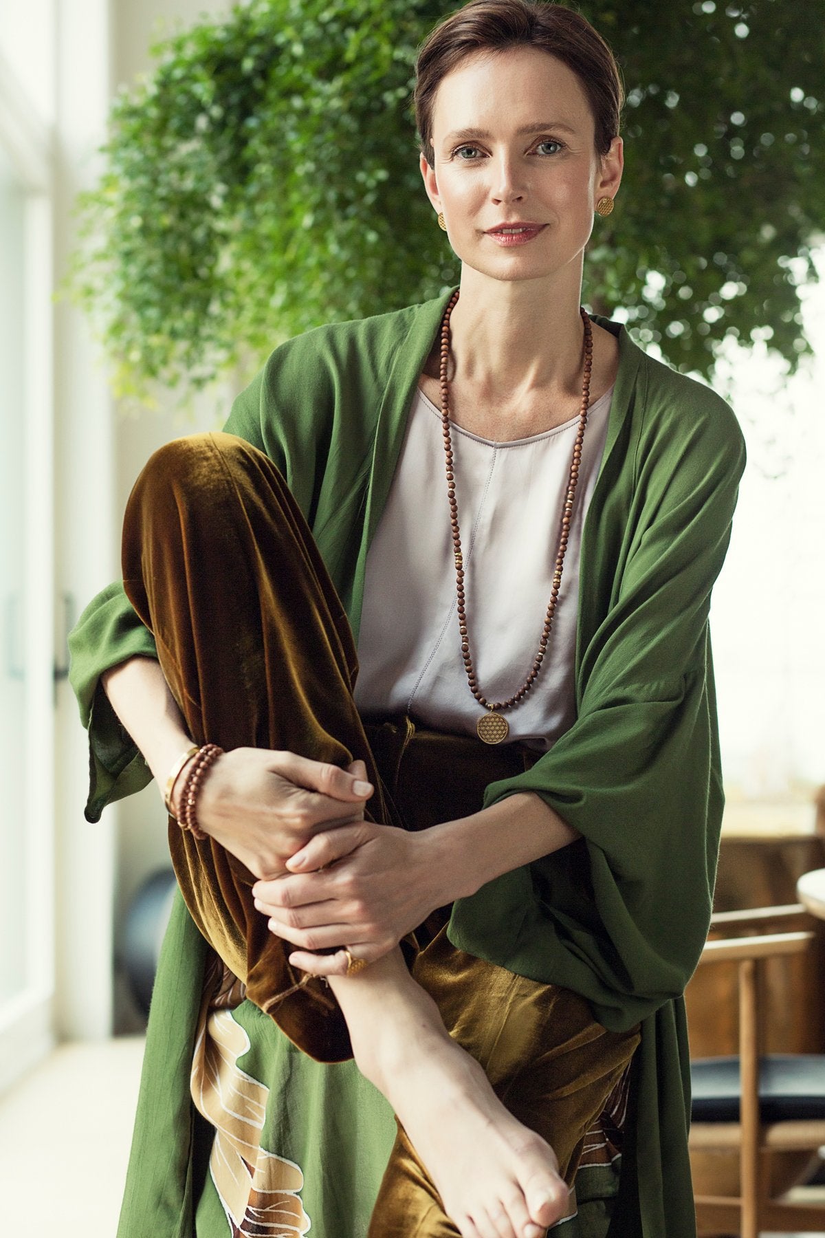 2. Woman in green robe wearing New Vintage by Kriss Flower Of Life gold stud earrings, seated in a bright room