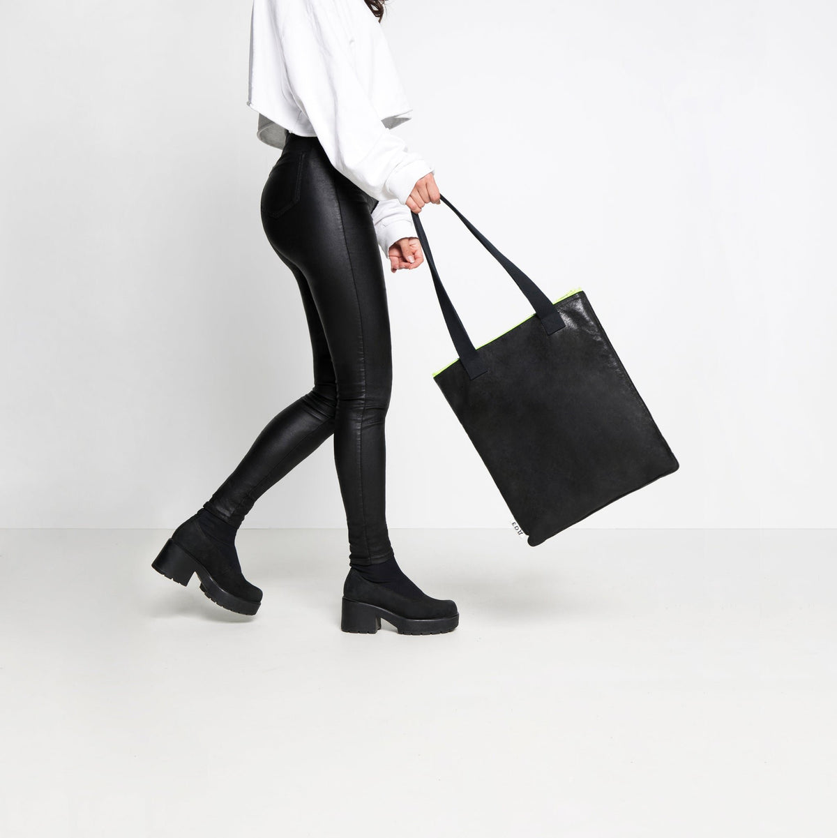 1. Woman carrying Daz Studio handmade black leather tote with neon green zipper, styled with black leggings and boots