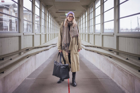 1. Woman in long beige coat and scarf holding black Stella Soomlais weekender bag in covered walkway