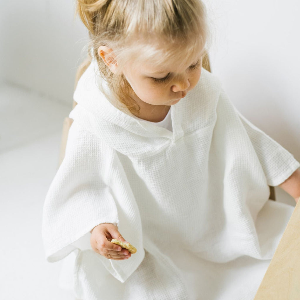 2. Child sitting in white linen waffle poncho holding a snack in a bright room